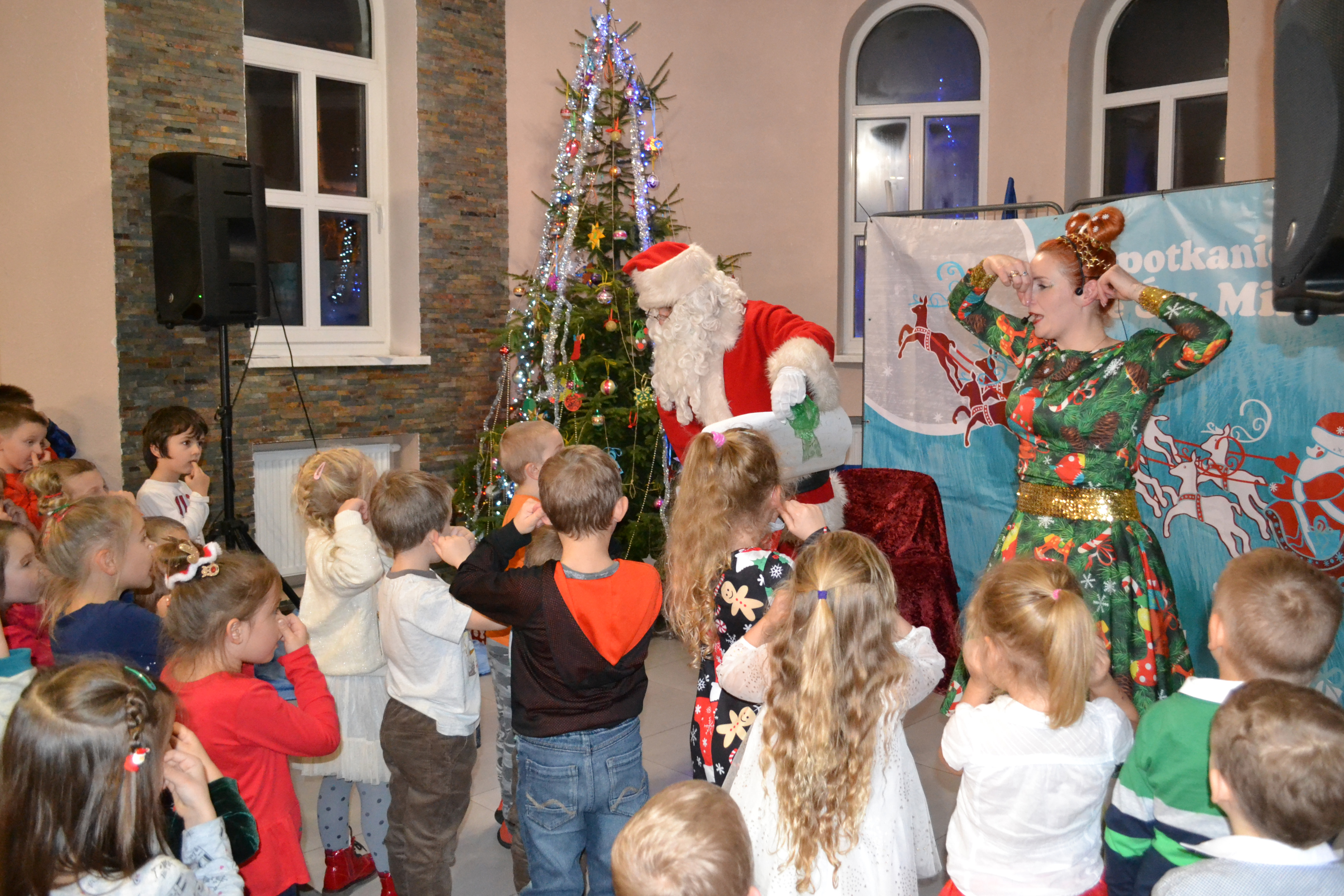 Spotkanie z Mikołajem w sali pod Goruszką, Święty Mikołaj stoi obok przystrojonej  choinki, wokół licznie zgromadzone dzieci