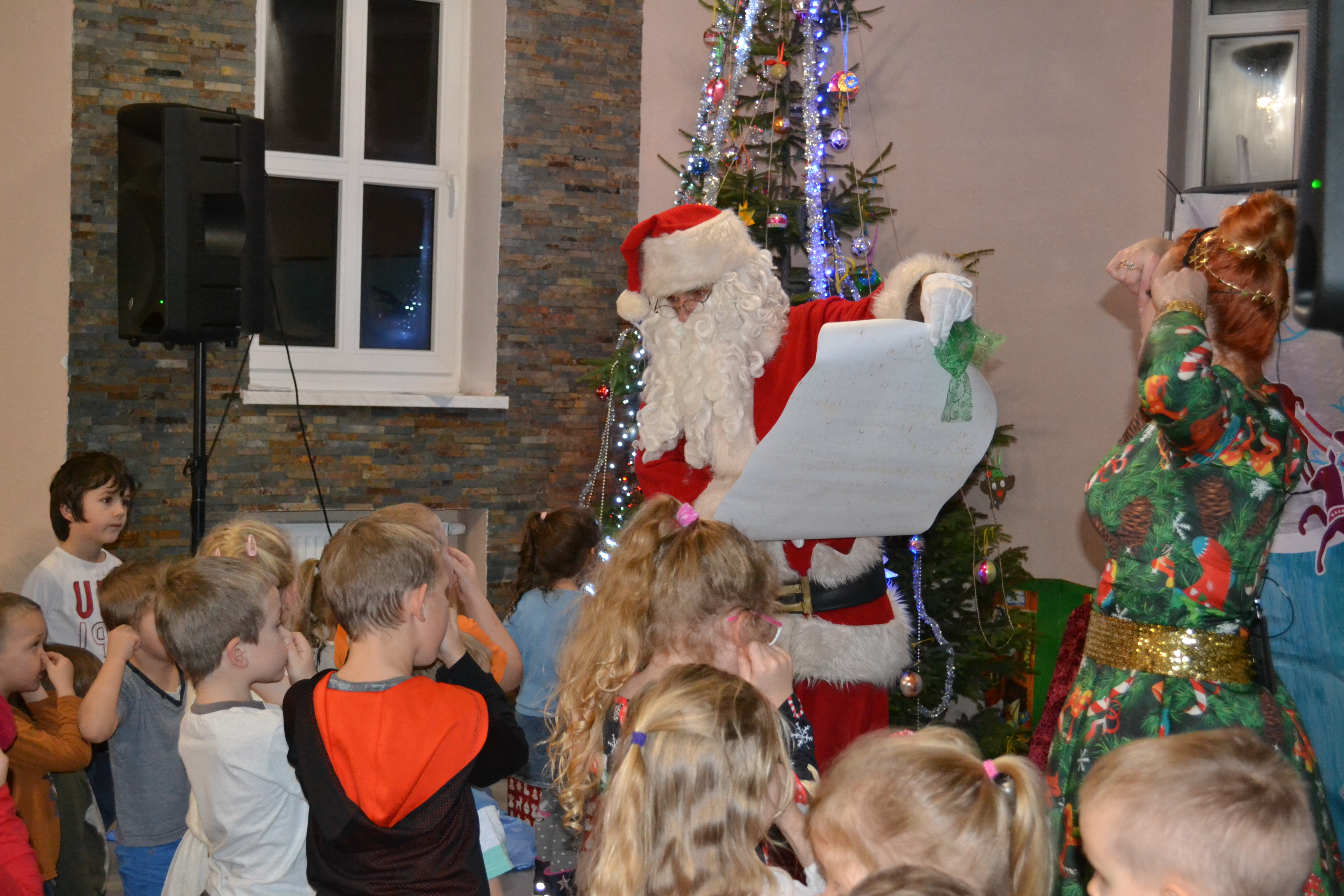 Spotkanie z Mikołajem w sali pod Goruszką, Święty Mikołaj i  Ciocia Tunia stoją na podwyższeniu, wokół licznie zgromadzone dzieci